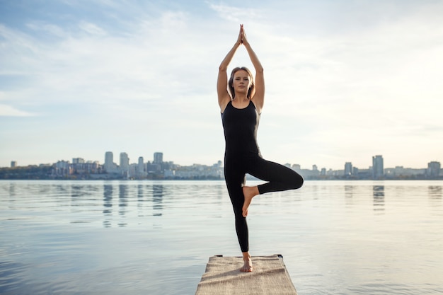 Esercitazione di yoga di pratica della giovane donna al molo di legno calmo con la città sport e ricreazione nella fretta della città