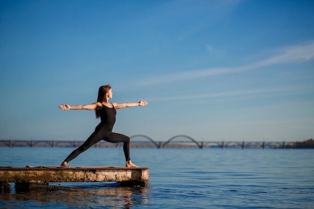 Young woman practicing yoga exercise at quiet wooden pier with city . Sport and recreation in city rush