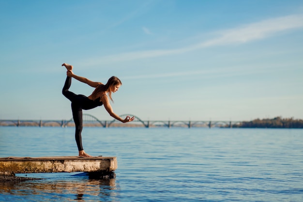 Young woman practicing yoga exercise at quiet wooden pier with city . Sport and recreation in city rush