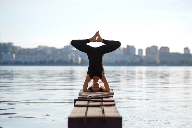 Young woman practicing yoga exercise at quiet wooden pier with city . Sport and recreation in city rush