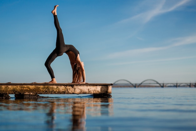 Young woman practicing yoga exercise at quiet wooden pier with city 