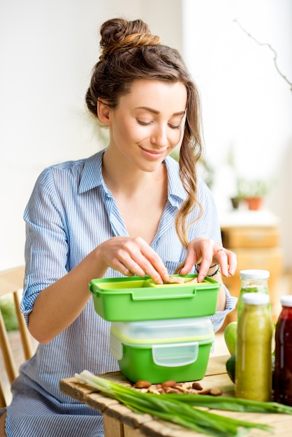 屋内のテーブルに座っている緑のお弁当箱で食べ物をprapairing若い女性