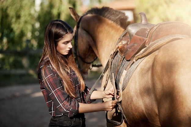 Young woman preparing to ride a horse tying down the saddle