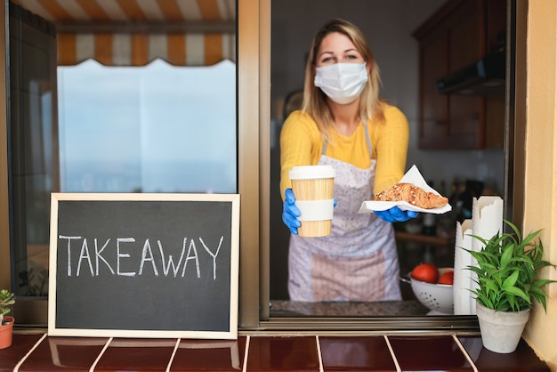 安全マスクを着用しながらパン屋でテイクアウトの朝食とコーヒーを準備する若い女性-食べ物に焦点を当てる