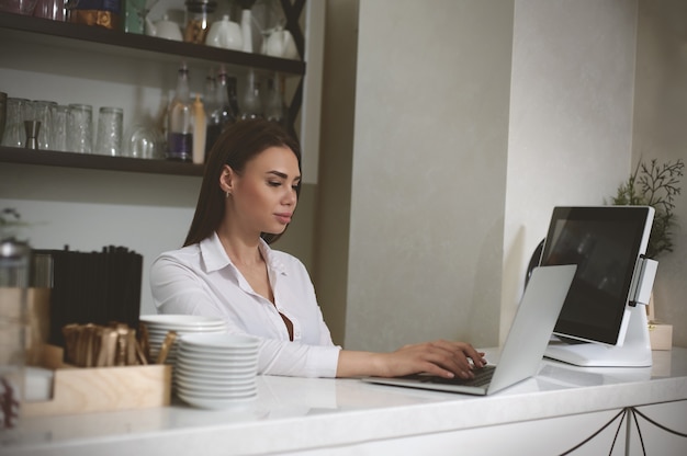 A young woman prints text on laptop standing behind a bar