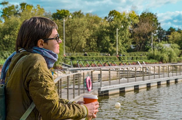 Young woman in profile with cup of coffee stands on the pier of city pond and looks at the water