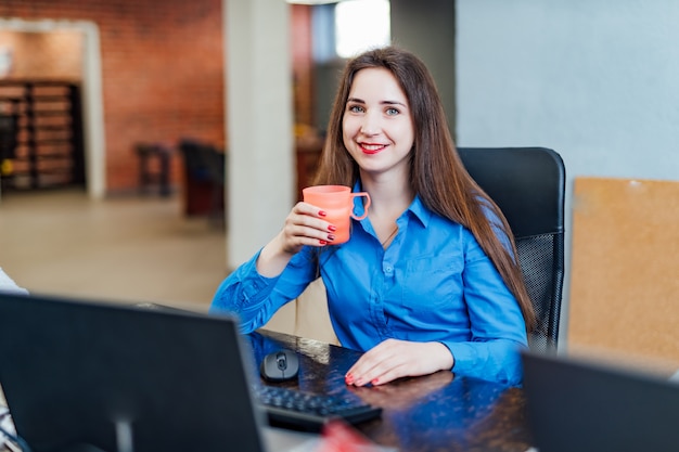 Programmatore della giovane donna che si siede in un companyfice nel computer anteriore con una tazza rosa. ingegnere informatico professionista che esamina macchina fotografica e sorridere. lavoro software