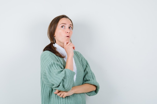 Young woman propping chin on hand, looking up in blouse, cardigan and looking wondered , front view.