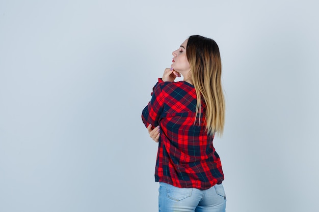 Young woman propping chin on hand, standing with her back in checked shirt, jeans and looking confident. front view.