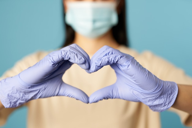young woman in protective face mask and gloves showing heart shape with hands