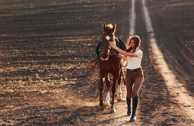 晴れた日中に農業分野で彼女の馬と一緒に歩く保護帽子をかぶった若い女性