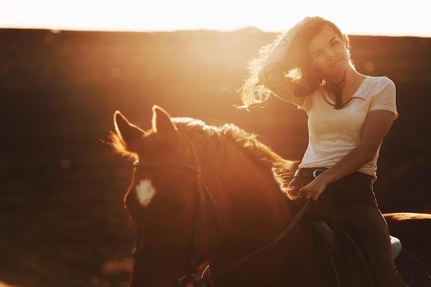 Young woman in protective hat with her horse in agriculture field at sunny daytime