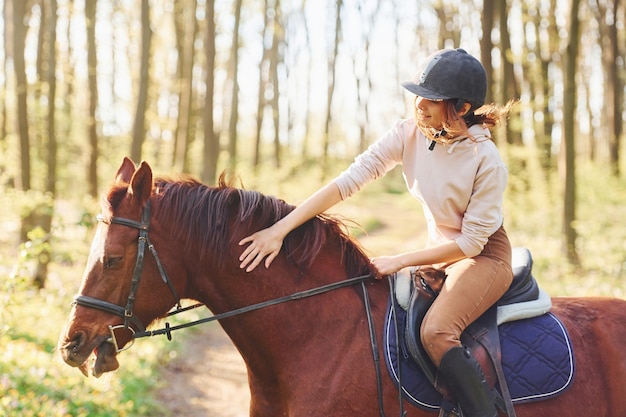 Young woman in protective hat with her horse in the forest at sunny daytime