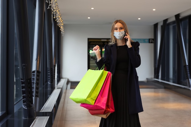 Молодая женщина в защитной маске после покупок разговаривает по телефону в торговом центре