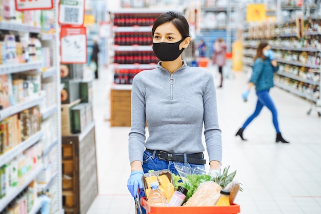 Giovane donna in maschera protettiva e guanti scegliendo prodotti al supermercato. acquisti sicuri durante una pandemia.