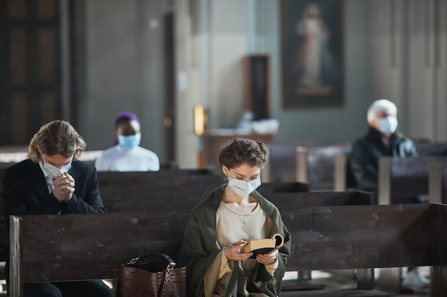 Foto giovane donna in maschera protettiva seduta su una panchina e leggendo la bibbia in chiesa