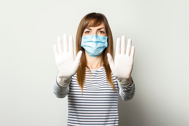 Foto la giovane donna in una mascherina medica protettiva e guanti in lattice tiene le mani davanti e mostra lo spettatore su un muro isolato luce. concetto coronavirus, pandemia, gesto mani pulite. copia spazio