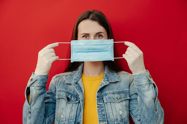 若い女性はマスクで顔を保護し、流行病の危険を冒していると考え、ウイルスに感染しており、赤い壁に隔離されたカメラを見ています。コンセプトの安全性、流行、コロナウイルス、ウイルス保護