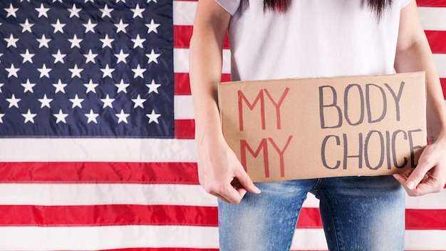 Young woman protester holds cardboard with My Body My Choice sign against USA flag