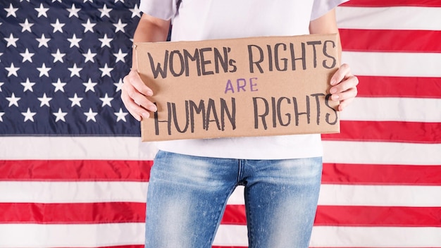 Young woman protester holds cardboard with Womens Rights Are Human Rights sign against USA flag