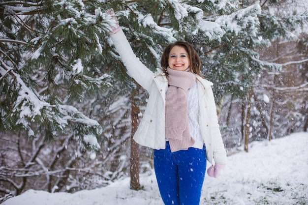 若い女性はトウヒの枝を引っ張って自分自身に雪を投げます暖かい服を着た女性は冬の公園で楽しんでいます