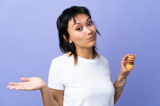 Young woman over purple wall holding colorful French macarons and making doubts gesture