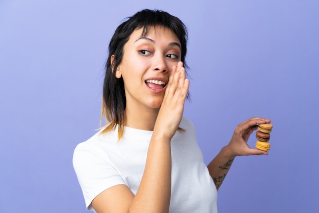 Young woman over purple wall holding colorful French macarons and whispering something