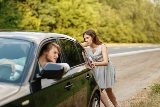 Young woman pushing broken car with man on road, breakdown