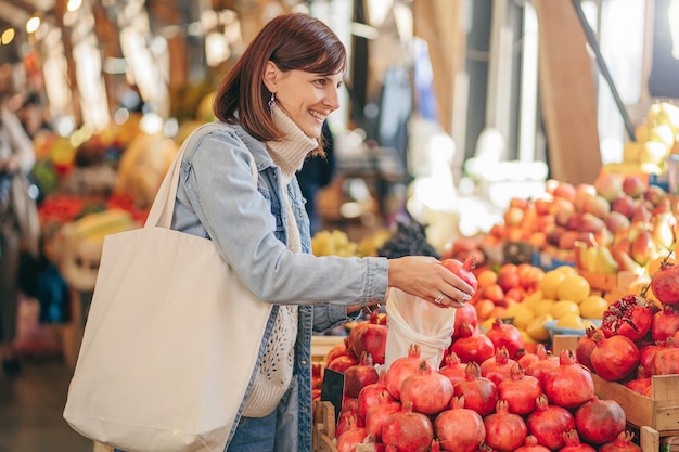 Молодая женщина кладет фрукты и овощи в мешок из-под хлопка на продуктовом рынке. Многоразовая эко-сумка для покупок. Устойчивый образ жизни. Экологичная концепция.