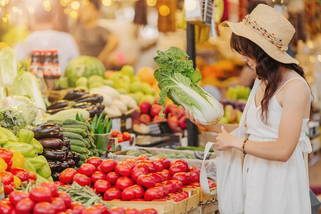 若い女性は、食品市場で綿花の農産物の袋に果物や野菜を入れます。ショッピング用の再利用可能なエコバッグ。ゼロウェイストのコンセプト。