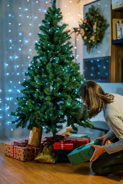 Foto giovane donna mette regali sotto un albero di natale con ghirlande luccicanti nella vista posteriore della stanza