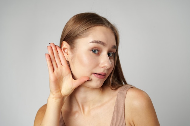 Young woman puts a hand to the ear to hear better on gray background