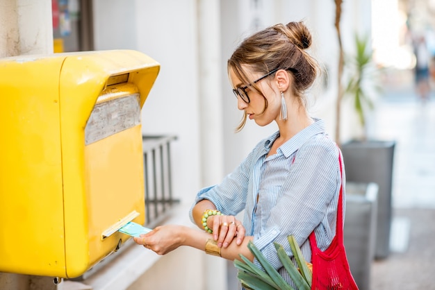 写真 通りの屋外に立っている古い黄色のメールボックスに手紙を置く若い女性