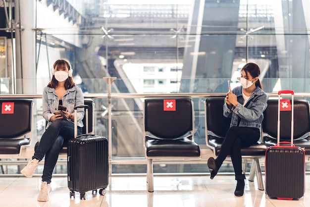 Young woman in quarantine for coronavirus wearing surgical mask face protection during virus pandemic with social distancing use smartphone typing message with luggage wait flight travel at airport