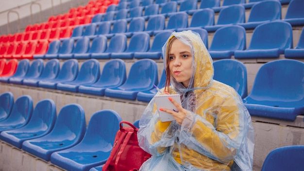 Young woman in raincoat with notepad pen sitting on stadium bleachers alone in rainy weather Female journalist writing down notes during sports training at street stadium