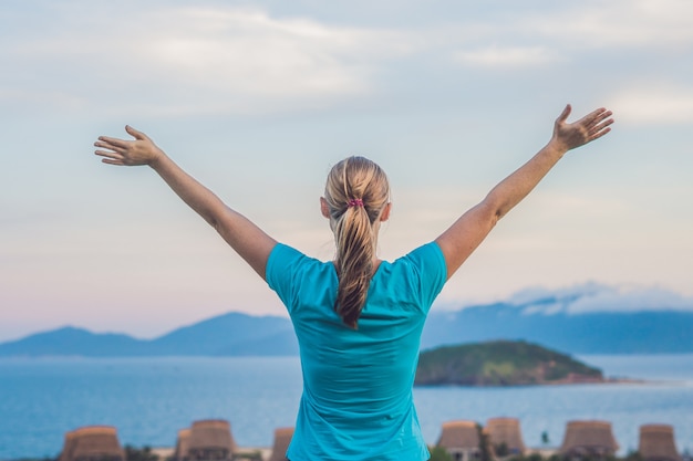 The young woman raised her hand up to the sun, against the sea, rocks and sky with clouds.