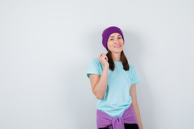 Young woman raising hand while posing in blue t-shirt, purple beanie and looking happy , front view.