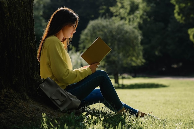 Молодая женщина читает книгу возле дерева в парке в солнечный день