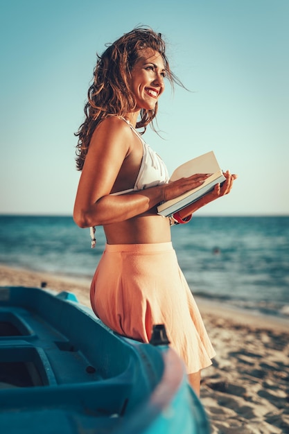 Young woman reading a book on the sandy beach and thinking with smile on her face.