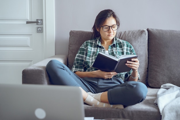 Young woman reading a book sitting on the sofa at home. distance work or online education. stay home during self-isolation