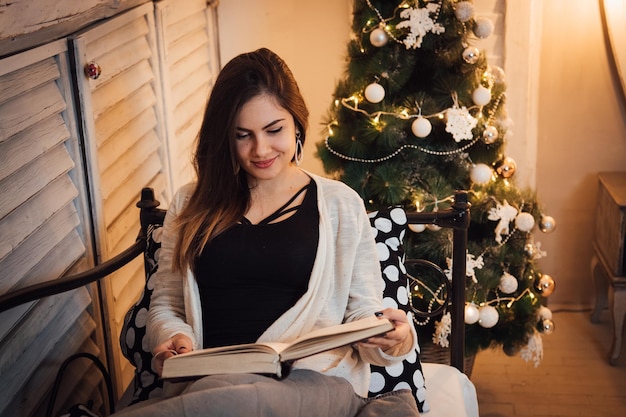 Young woman reading book while sitting on sofa by christmas tree at home