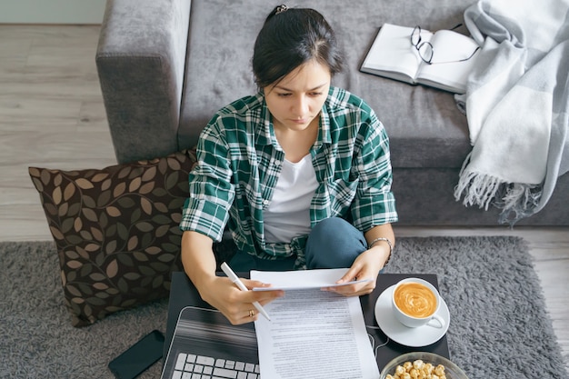 Young woman reading documents while working on a laptop at home