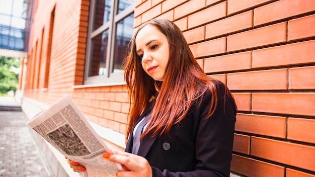 Young woman reading newspaper on street Side view of young woman with long hair in casual clothes standing on street and reading newspaper