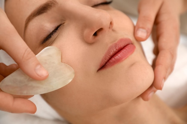 Young woman receiving facial massage with gua sha tool in beauty salon closeup