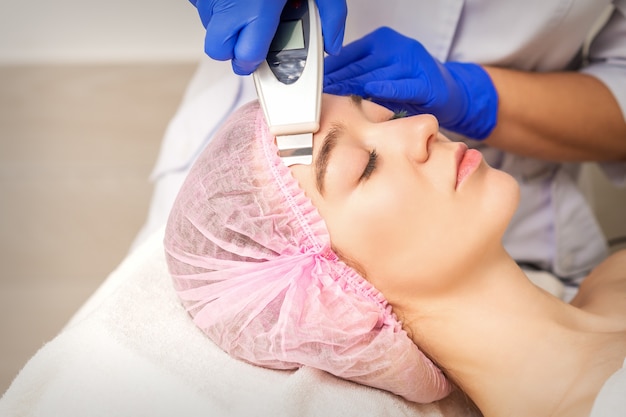 Young woman receiving facial skin cleaning by ultrasonic cosmetology face equipment in medical salon