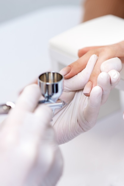 Young woman receiving manicure by airbrush in nail salon. Procedure for spraying paint on the nails