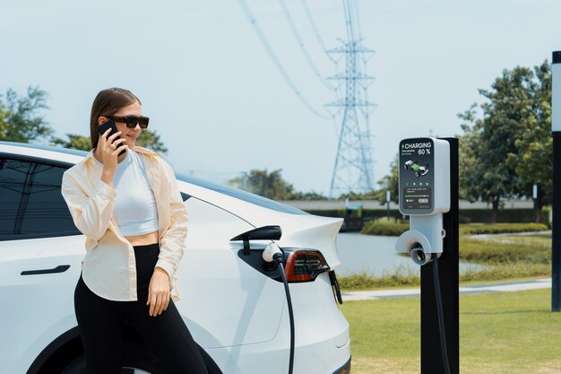 Photo young woman recharging ev car battery while talk on phone at charging station connected to electrical power grid tower facility as electrical industry for eco friendly vehicle utilization expedient