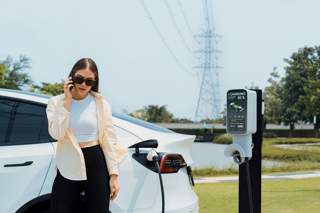 電動車のバッテリーを充電しながら電話で話す若い女性 電気グリッドタワー施設に接続された電気産業として エコフレンドリーな車両の利用に適しています