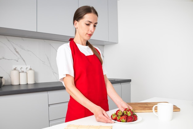 キッチンでイチゴのプレートを保持している赤いエプロンの若い女性
