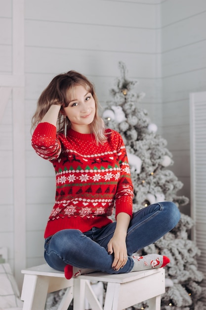 A young woman in a red Christmas sweater with deer pattern siitting on the background of a fir tree